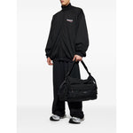 Model wearing black Balenciaga Coats Black blouson jacket with front zip and side pockets, holding a black duffle bag.
