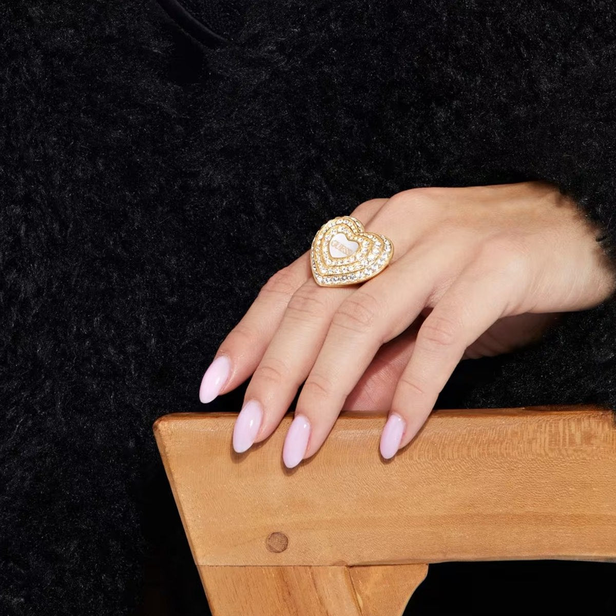 Close-up of a hand wearing GUESS JEWELS JEWELRY Mod. JUBR04016JWYGWH54 heart-shaped gold ring on a wooden chair.
